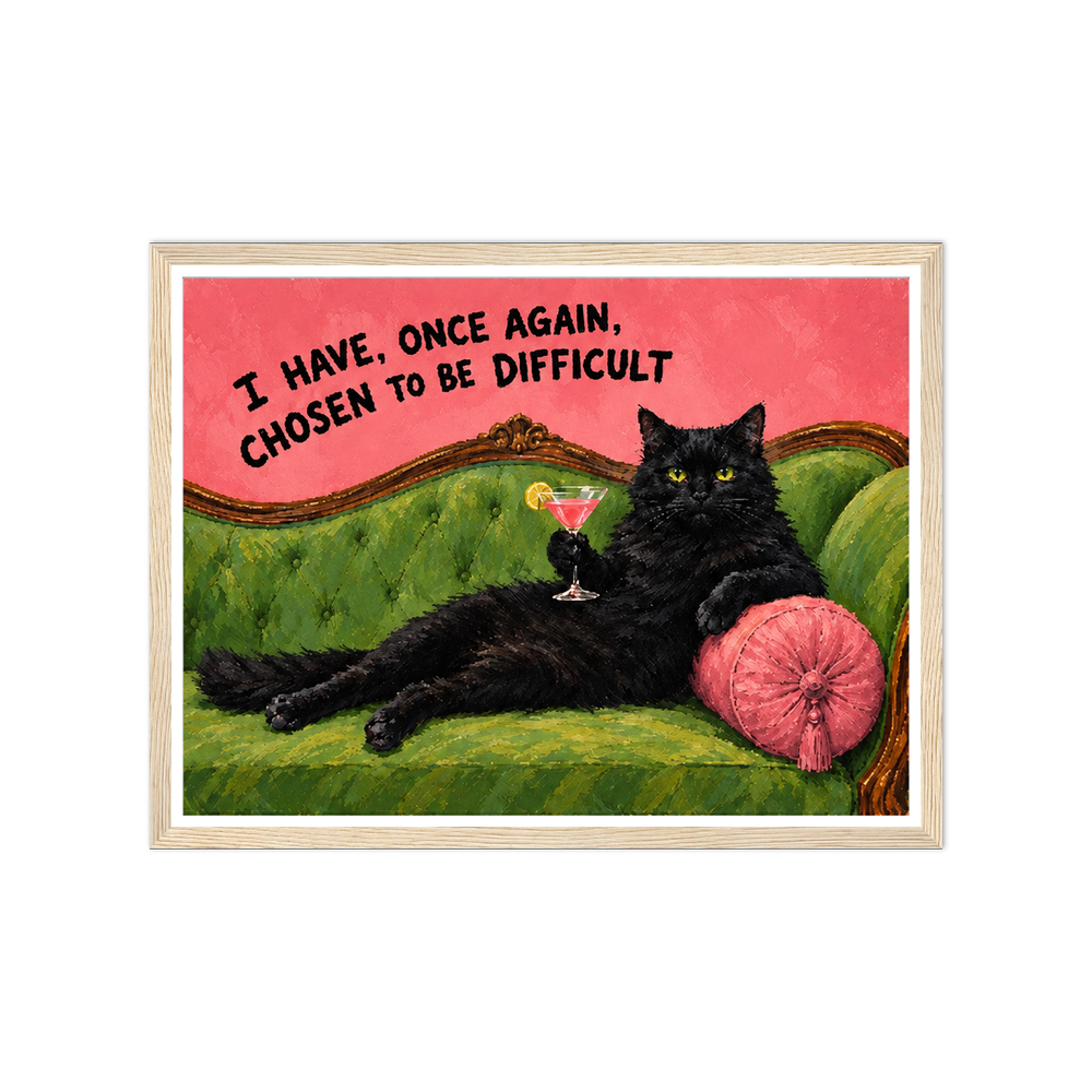 Black cat with a cocktail on a green sofa beneath a chosen to be difficult quote in a natural wood frame.