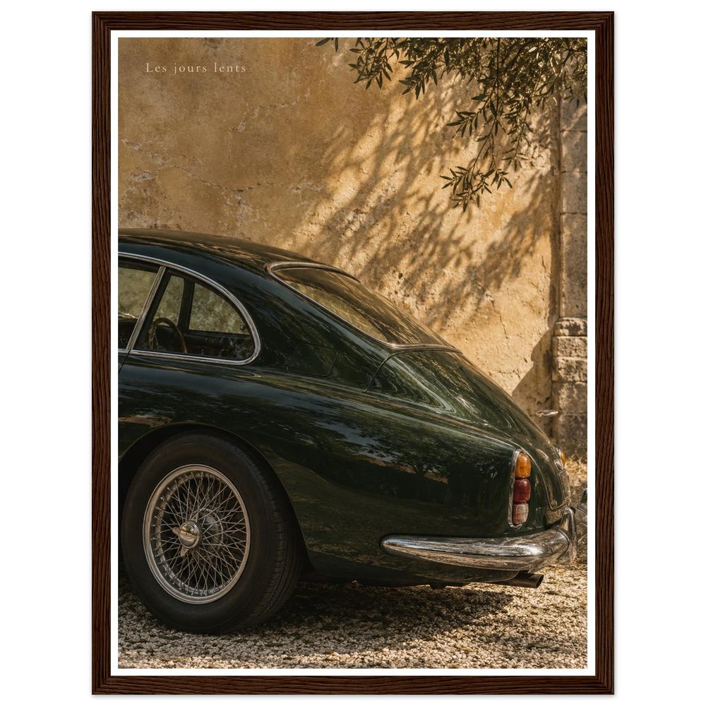 Deep green classic car beside a sunlit ochre wall with leafy shadows in a dark wood frame.